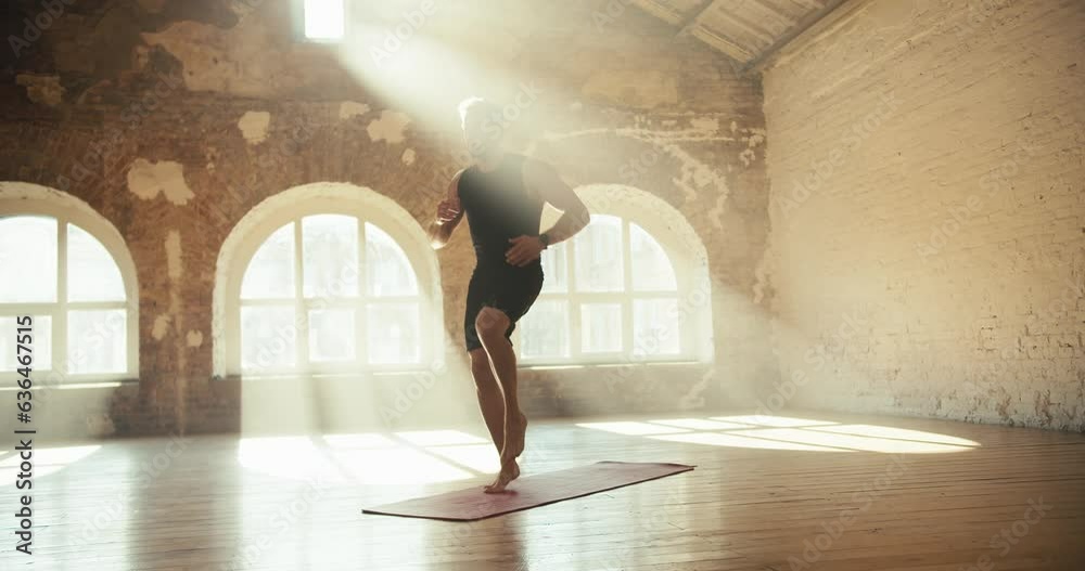 A blond man in a black sports uniform does exercises and runs in place on a special rug in a sunny hall with brick walls. Video filmed in high quality