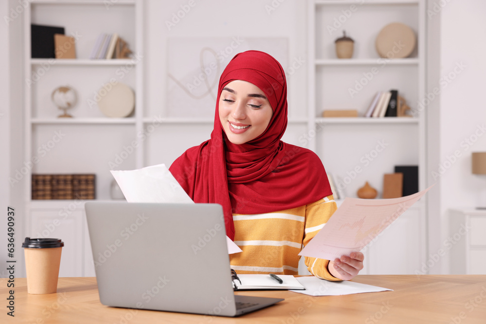 Muslim woman working near laptop at wooden table in room Muslim woman working near laptop at wooden table in room