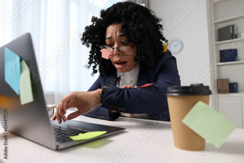 Deadline concept. Stressed woman checking time near laptop in office. Many sticky notes everywhere as reminders