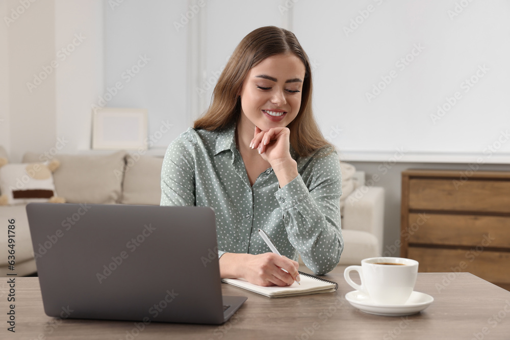 Happy woman writing something in notebook near laptop at wooden table in room Happy woman writing something in notebook near laptop at wooden table in room