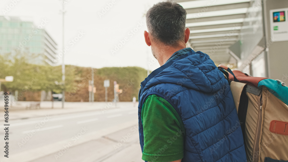 Middle-aged man dressed in casual clothes, with backpack on his shoulder, looks around while waiting for tram at public transport stop in the city Middle-aged man dressed in casual clothes, with backpack on his shoulder, looks around while waiting for tram at public transport stop in the city
