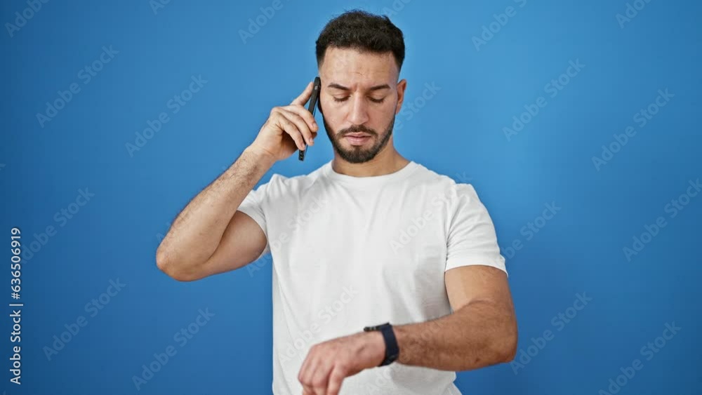 Young arab man talking on smartphone looking watch walking away over isolated blue background
