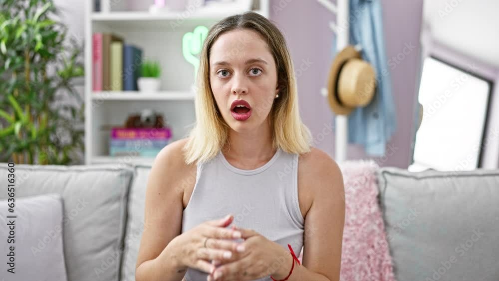 Young blonde woman sitting on sofa surprised covering mouth with hands at home Young blonde woman sitting on sofa surprised covering mouth with hands at home