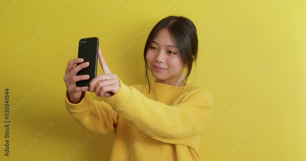 Young Beautiful Asian woman holding mobile phone taking photo, selfie Happy girl teenager enjoy taking smile selfie with smartphone express exited and positive emotion Isolated yellow background Young Beautiful Asian woman holding mobile phone taking photo, selfie Happy girl teenager enjoy taking smile selfie with smartphone express exited and positive emotion Isolated yellow background