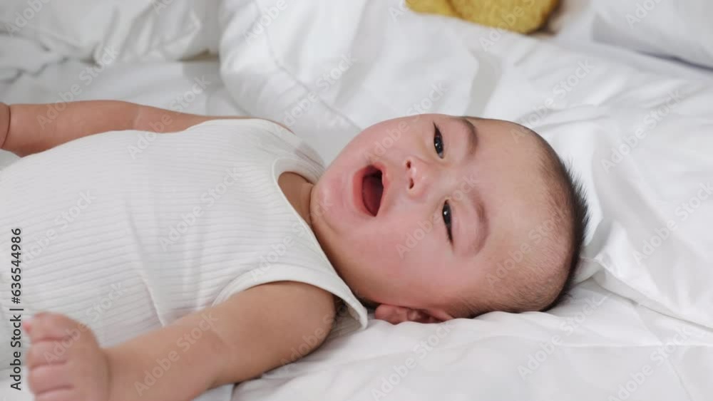 Asian smiling infant child at home, Cute little baby on bed under soft blanket, baby wearing bodysuit lying on white beedsheets in bedroom Asian smiling infant child at home, Cute little baby on bed under soft blanket, baby wearing bodysuit lying on white beedsheets in bedroom