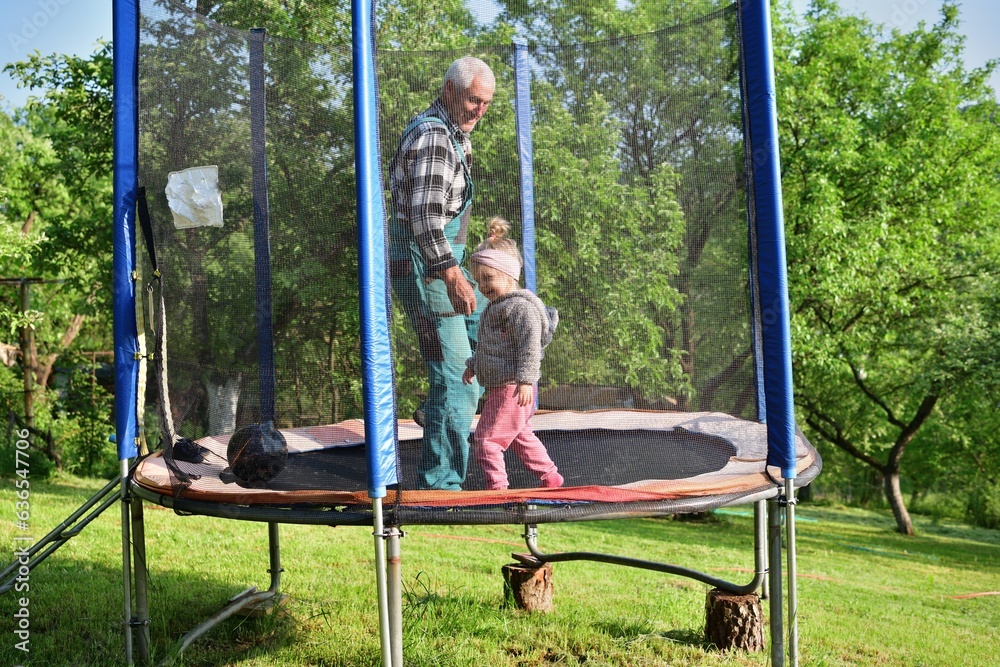 A little girl runs around in a trampoline and kicks a ball with her grandfather