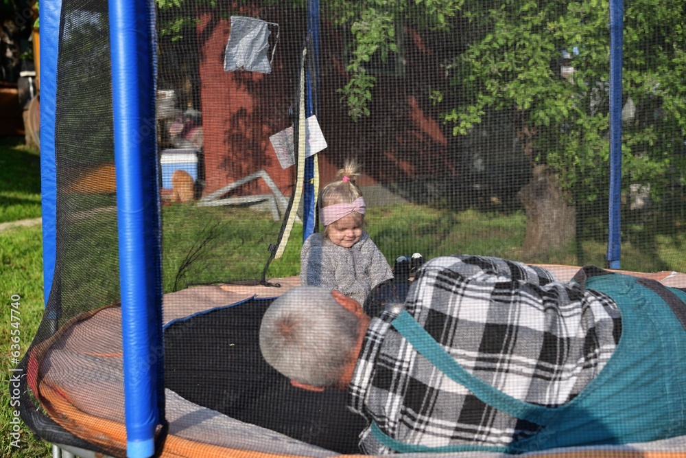 A little girl runs around in a trampoline and kicks a ball with her grandfather