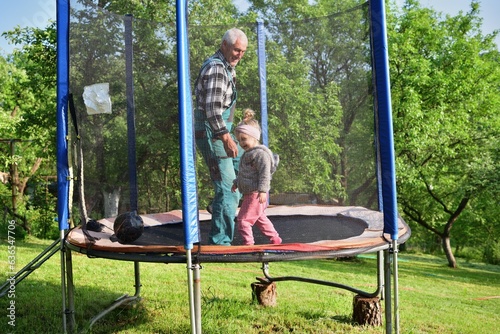 A little girl runs around in a trampoline and kicks a ball with her grandfather