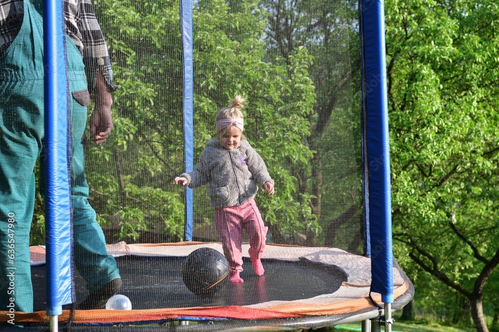 A 2-year-old girl jumps and kicks a ball on a trampoline in the summer