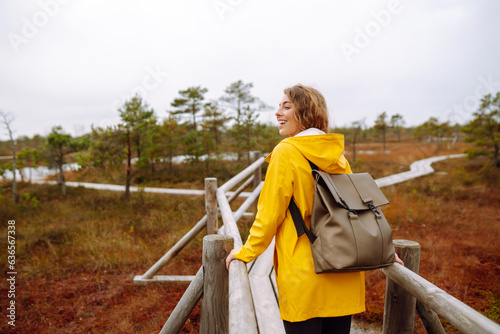 Cheerful woman in a yellow coat walks, explores a wooden path of wetlands. The concept of travel, vacation, nature. Active lifestyle.