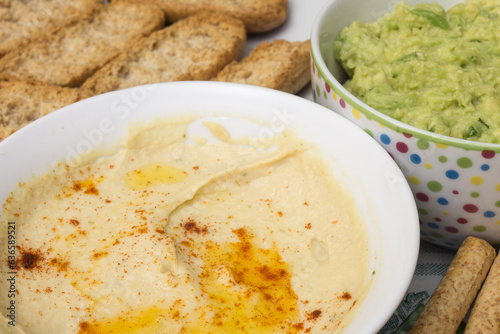 A close-up shot of a bowl of hummus dip, made with chickpeas, tahini, garlic and lemon. The background is blurred, but you can see a bowl of guacamole, whole wheat toast and bread sticks.