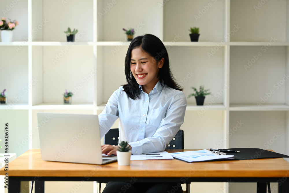 Charming female entrepreneur in blue shirt using laptop computer, doing online data market analysis at office Charming female entrepreneur in blue shirt using laptop computer, doing online data market analysis at office