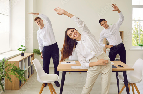 Fototapeta Naklejka Na Ścianę i Meble -  Happy young business people doing stretching sport exercises standing at the workplace in modern office. Group of smiling company employees exercising fitness for health during a break from a work.