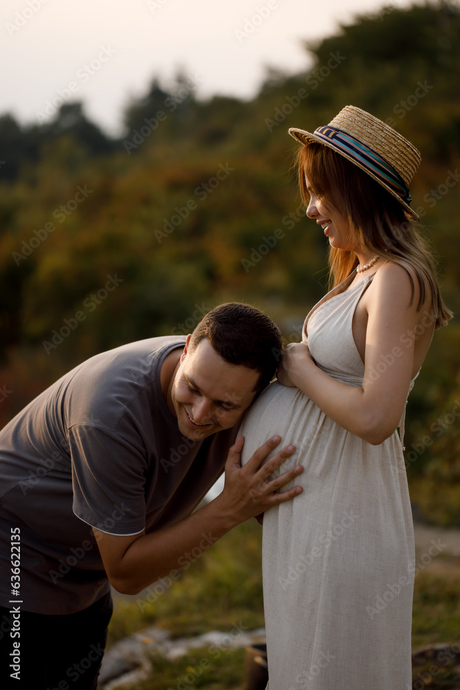 A young couple in love are standing against the background of the sunset, looking at each other, smiling. A woman holds a pregnant belly in her hands. A young couple in love are standing against the background of the sunset, looking at each other, smiling. A woman holds a pregnant belly in her hands.