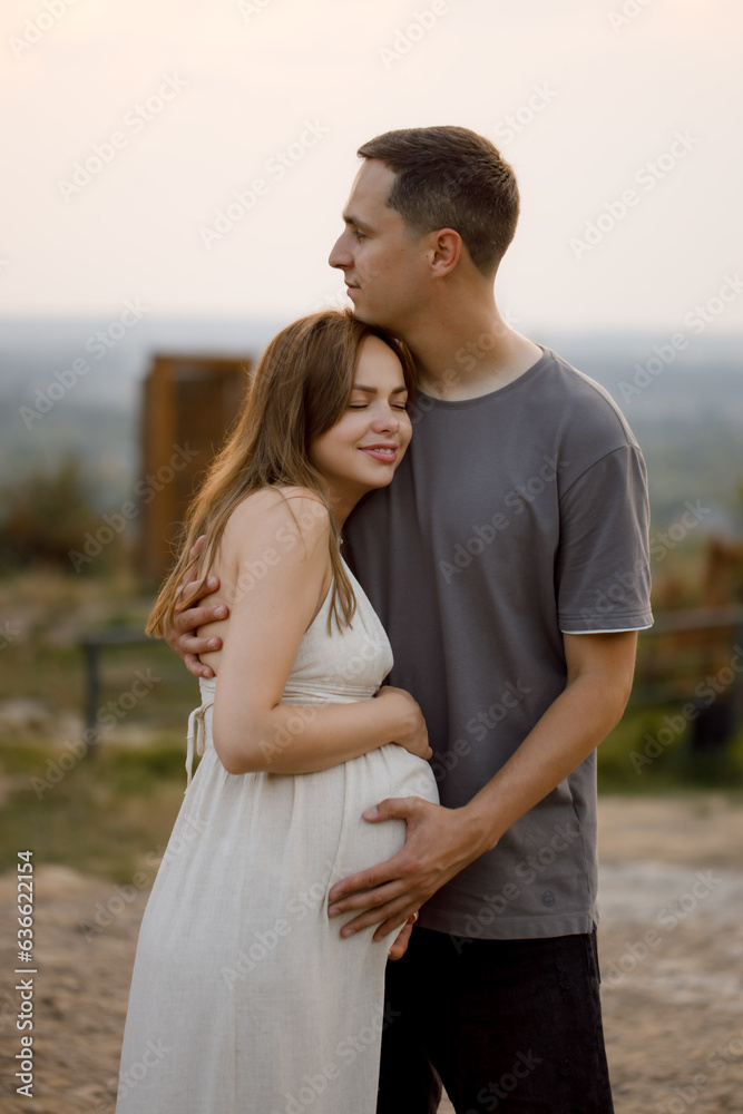 A young couple in love are standing against the background of the sunset, hugging, kissing and holding a heavy belly in their hands.