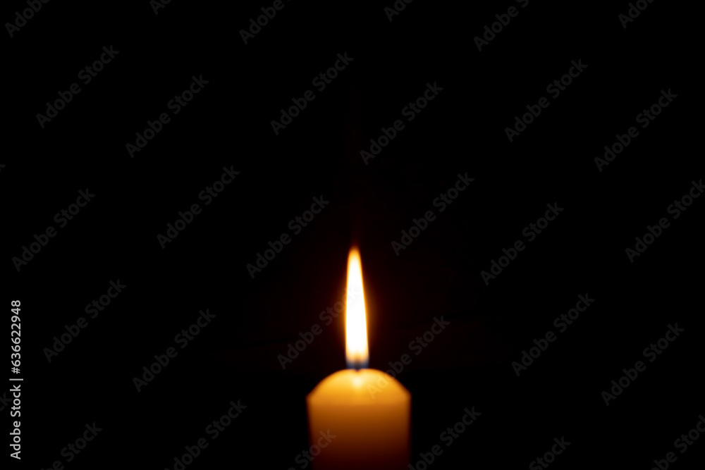 Blurred photo of single burning candle flame or light glowing on a big yellow candle on black or dark background on table in church for Christmas, funeral or memorial service with copy space. Blurred photo of single burning candle flame or light glowing on a big yellow candle on black or dark background on table in church for Christmas, funeral or memorial service with copy space.