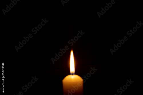 Blurred photo of single burning candle flame or light glowing on a big yellow candle on black or dark background on table in church for Christmas, funeral or memorial service with copy space.
