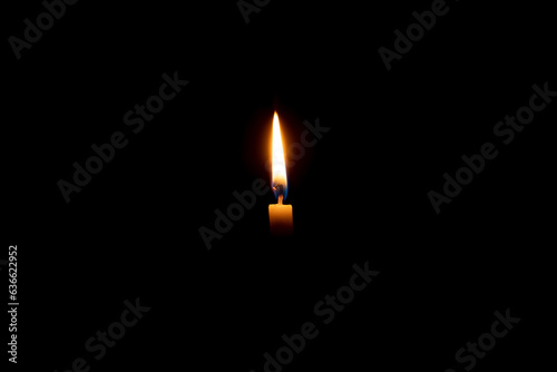 Single burning candle flame or light glowing on a small white candle on black or dark background on table in church for Christmas, funeral or memorial service with copy space.
