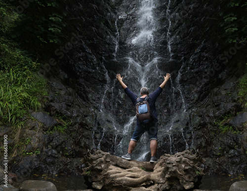 back, rear view of Tourists Man with backpack standing. Hands up on background of Waterfall in Mirveti, Adjara, Georgia. Summertime season. photo