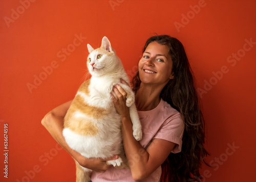Happy young brunette caucasian woman holding her white and red cat on orange background.