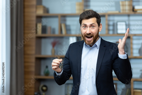 Portrait of a happy and successful realtor broker, the man looks joyfully at the camera and holds the keys to a new apartment in the house, a businessman in a business suit works inside the office.