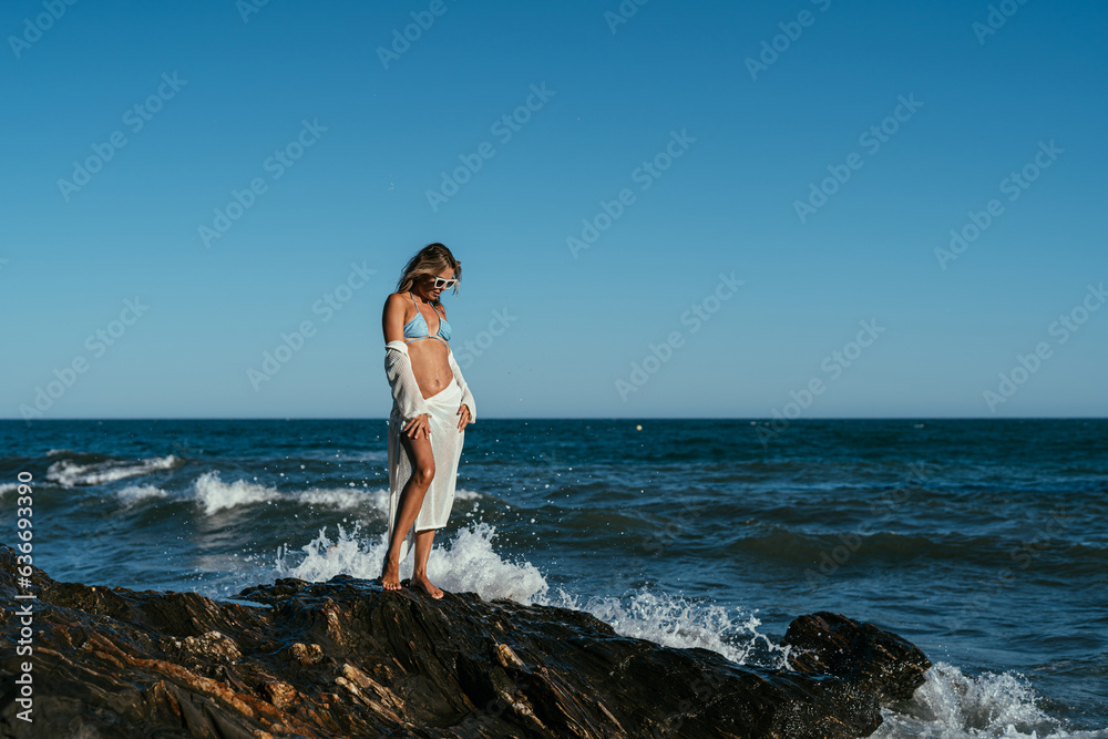 A beautiful girl in white glasses and white clothes sits on the rocks on the seashore