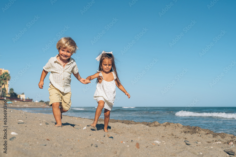 a little blond boy and a little brunette girl run on the beach on the sand in front of the sea on a sunny summer day a little blond boy and a little brunette girl run on the beach on the sand in front of the sea on a sunny summer day