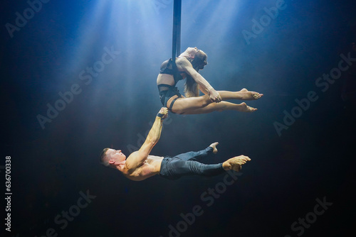 Aerial straps duo performance: a man and woman execute graceful acrobatic feats in mid-air against a black background, dressed in black and illuminated by a white-blue glow