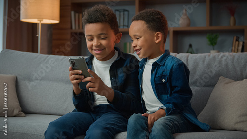 Children gadget addiction two happy laughing African American little boys brothers browsing mobile phone watching cartoons online without parental control at home couch kids siblings using smartphone