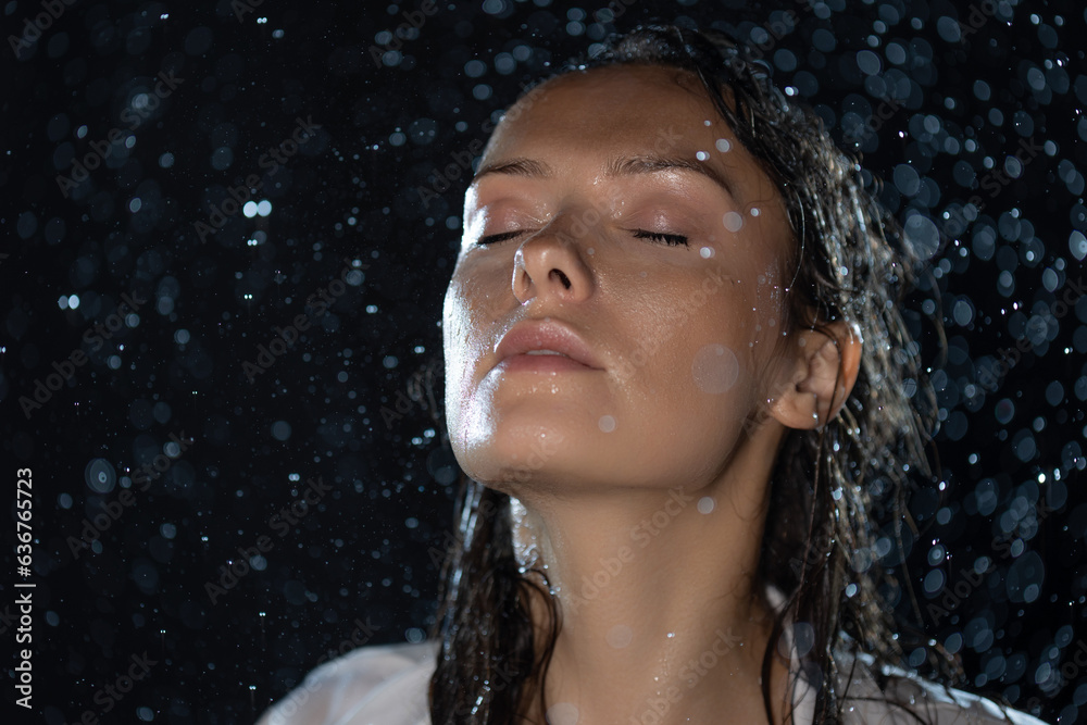 Fototapeta premium young woman in the rain, getting wet through, wet clothes and water running down her face. a beautiful brunette stands with her face exposed to the streams of water, portrait in aqua studio