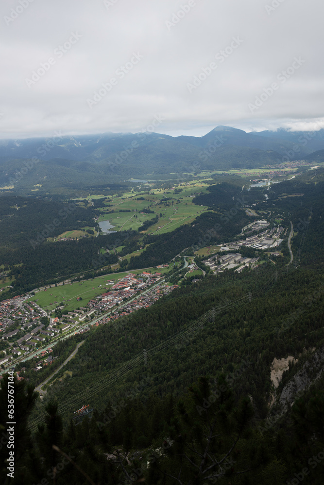 Fototapeta premium Aerial view of Mittenwald town surrounded by dense forest, green fields, and distant mountain ridge, captured from the Mittenwald High Trail under a cloudy sky.