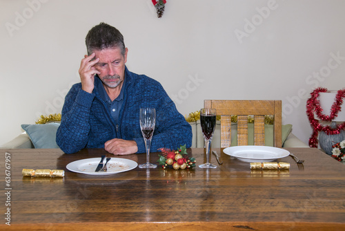 Loneliness at Christmas image. A man sits alone at a Christmas table with an empty plate by his side where someone significant would have been sat. Loss and solitude image. Selective focus on his face