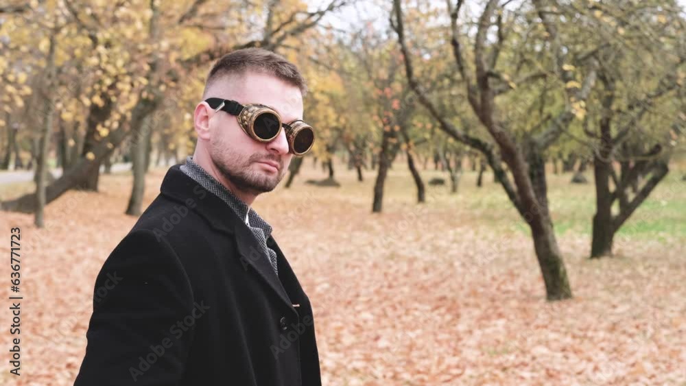 A man in a business suit, coat and steampunk goggles poses in an autumn park. Horizontal slow motion video A man in a business suit, coat and steampunk goggles poses in an autumn park. Horizontal slow motion video