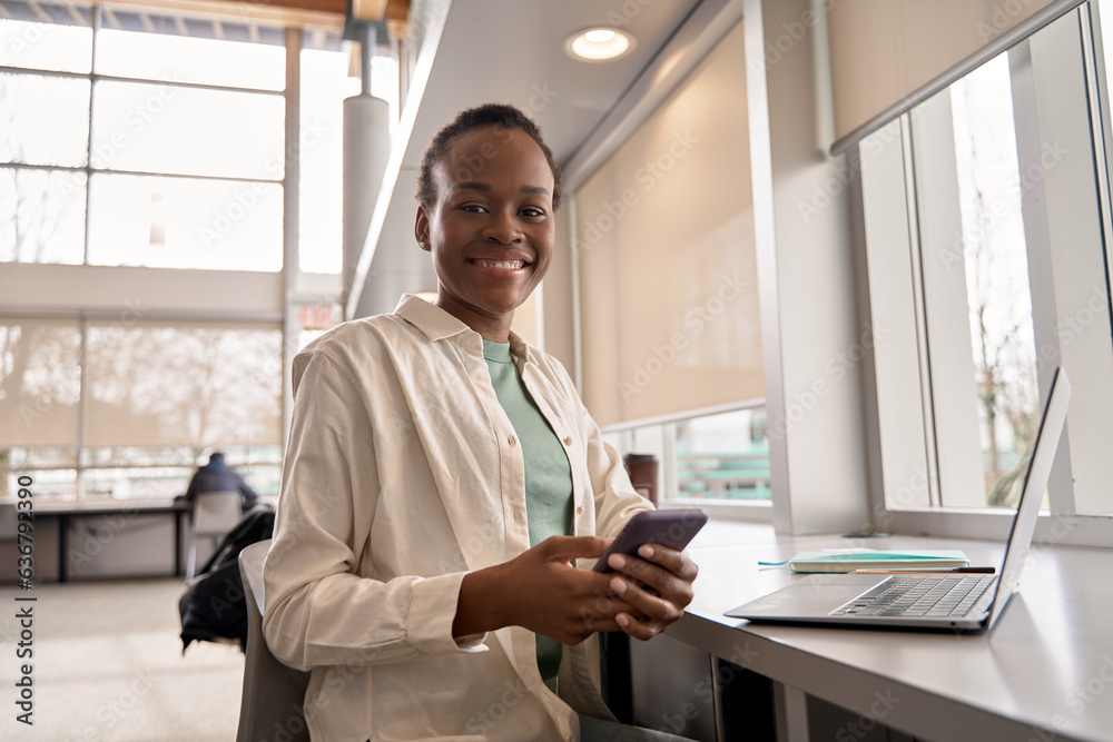 Happy African ethnic girl using mobile cell phone and laptop sitting at desk. Smiling Black teen student looking at camera holding smartphone tech elearning online in apps in university. Portrait.