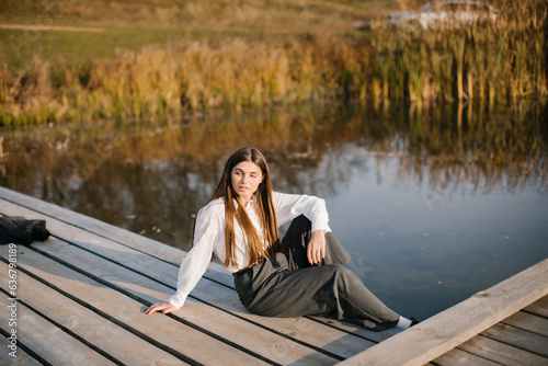 A woman with long hair in a white shirt and grey trousers is sitting on a wooden masonry near the lake in autumn.  © VikaNorm