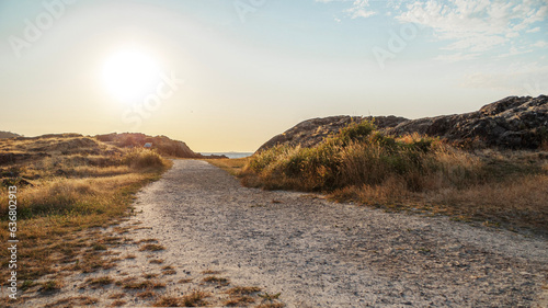 a gravel path winding through a rocky landscape with the sun setting in the background. The light-colored gravel path is bordered by grass and shrubs, and the rocky landscape features large boulders a