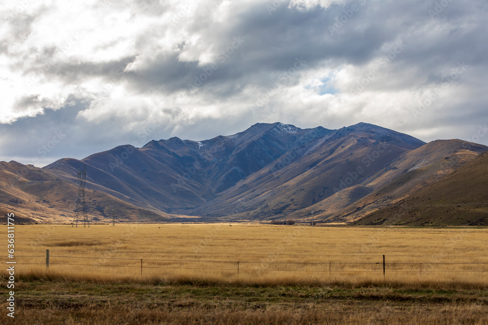 Fototapeta premium Photograph of a dry mountain range running behind a large brown agricultural field with low level grey clouds on the South Island of New Zealand