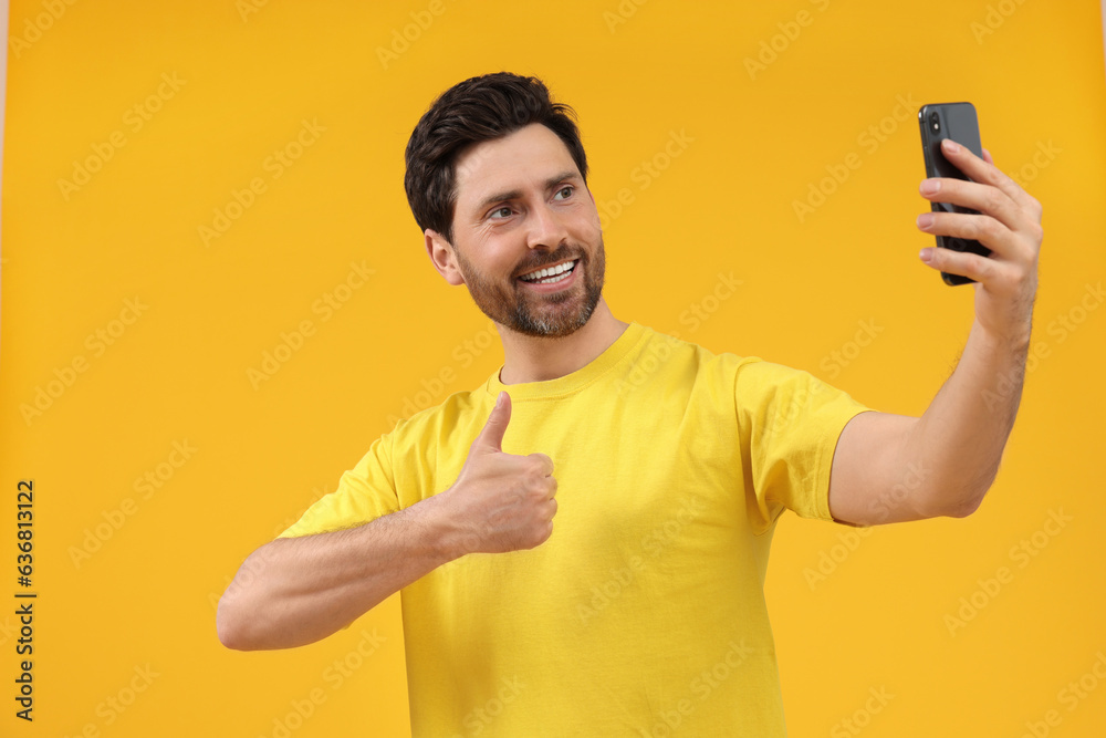 Smiling man taking selfie with smartphone and showing thumbs up on yellow background Smiling man taking selfie with smartphone and showing thumbs up on yellow background