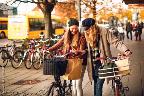 Young lesbian couple using a smart phone while shopping and pushing their bicycles on a city sidewalk