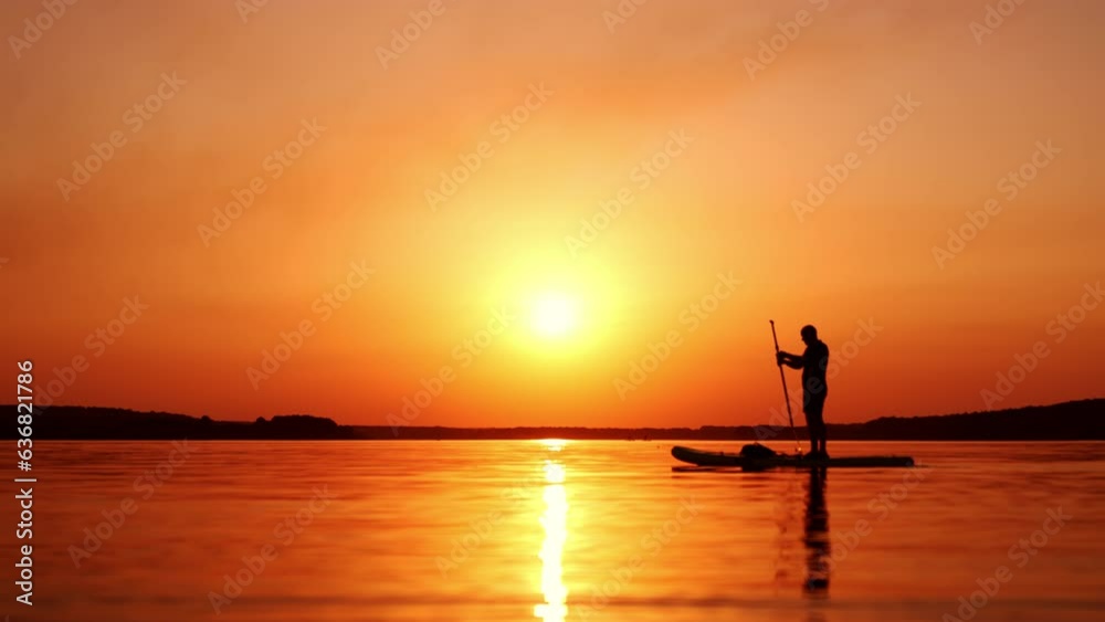Orange sunset over the river. Silhouette of a male standing on the sup board and slowly rowing with a paddle.