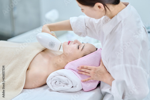 아시아 한국의 젊은 여성이 스파 또는 피부관리전문점 또는 피부과 병원에서 분홍색 두건을 쓰고 침대에 누워 베이지색 대형 타올을 덮고 하얀색 셔츠를 입은 전문관리사가 레이저 제모기로 얼굴부분 제모를 하고있다. 