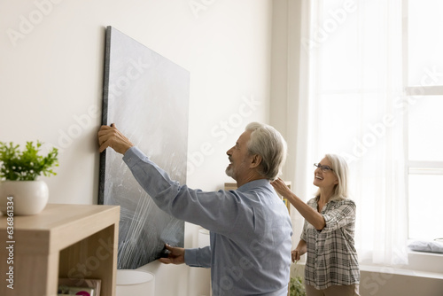Cheerful senior couple decorating apartment, hanging large picture on white wall, laughing, having fun, working on home interior contemporary design, enjoying domestic activities in new house
