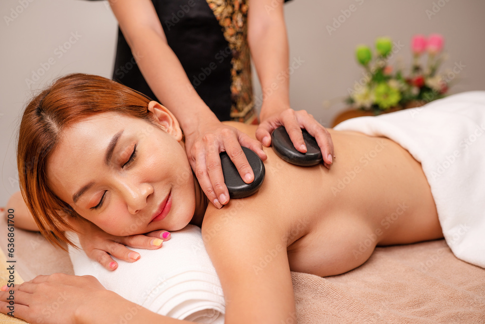 Woman enjoying exotic hot stones spa massage. Relaxed young woman lying on a spa bed while the masseuse is putting hot stones on her back, Spa treatment concept.