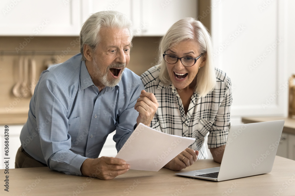 Joyful senior husband and wife reading legal document, paper report, letter at laptop, shouting for joy, making winner yes hand, celebrating financial success, investment income, profit, laughing Joyful senior husband and wife reading legal document, paper report, letter at laptop, shouting for joy, making winner yes hand, celebrating financial success, investment income, profit, laughing