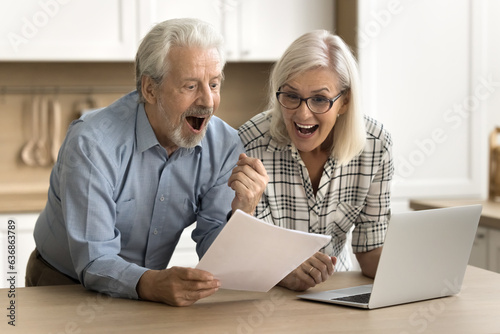 Joyful senior husband and wife reading legal document, paper report, letter at laptop, shouting for joy, making winner yes hand, celebrating financial success, investment income, profit, laughing