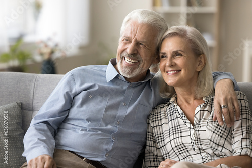 Cheerful old retired couple husband and wife enjoying leisure on home couch together, hugging, laughing, smiling, looking away, thinking on retirement, vacation plans, dreaming