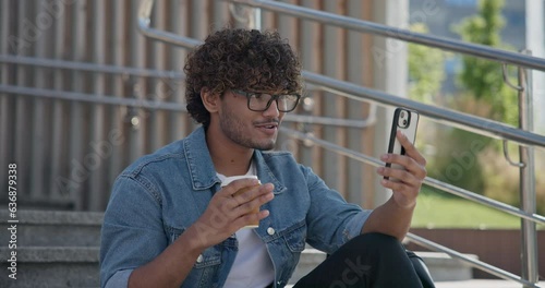 Young indian guy student sitting on a stairs, drinking coffee outdoors and talkink on the phone photo