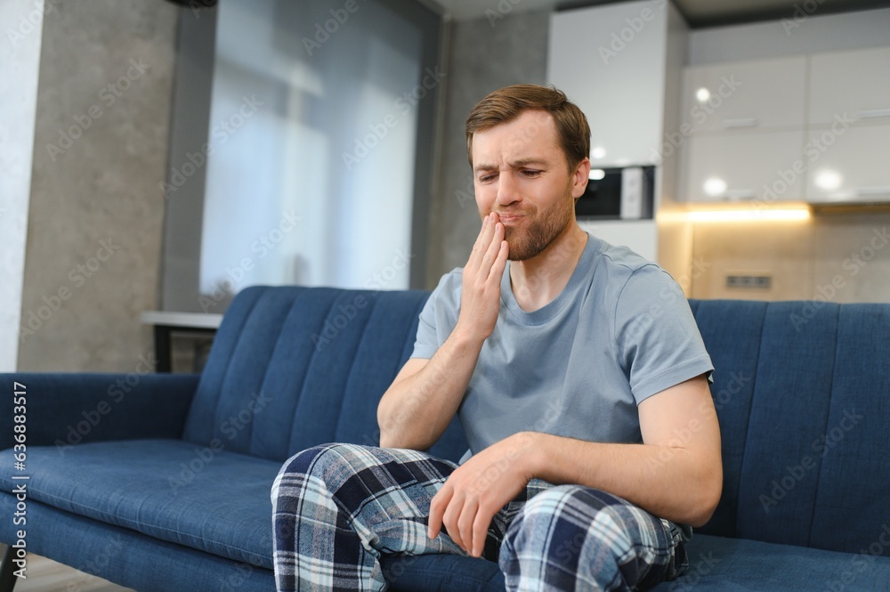 Toothache. Unhappy upset caucasian sad man sits on the couch at home, holds his hand near his cheek, grimaces, has an acute toothache, needs a consultation with a dentist doctor Toothache. Unhappy upset caucasian sad man sits on the couch at home, holds his hand near his cheek, grimaces, has an acute toothache, needs a consultation with a dentist doctor