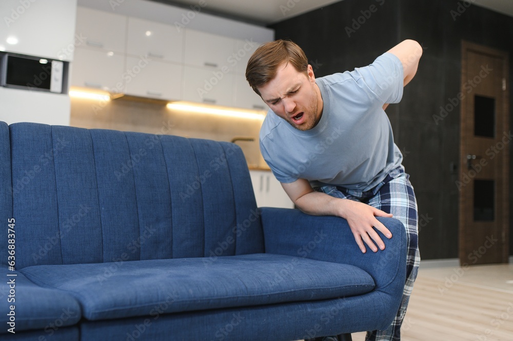 People, Healthcare and Treatment. Portrait of sick man suffering from acute side back pain, sitting on couch at home, person touching lower back aching area with hand. Health Problem Concept People, Healthcare and Treatment. Portrait of sick man suffering from acute side back pain, sitting on couch at home, person touching lower back aching area with hand. Health Problem Concept