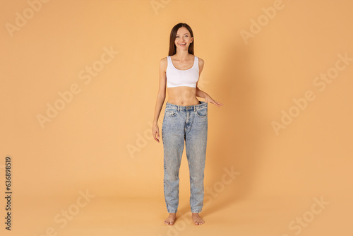 Diet, slimming concept. Happy young european lady wearing big jeans after weight loss diet program, posing on beige background, full length copy space